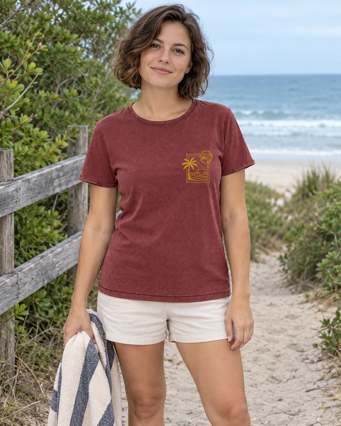 T-shirt vieilli femme rouge grenat délavé avec un timbre palmier 971 avec mer et soleil imprimé en jaune moutarde, porté par une femme dans un studio à la lumière chaude